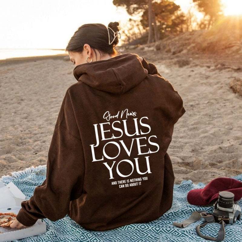 Person wearing a brown Kangaroo Pocket Hoodie with "Jesus Loves You" text on the back, sitting outdoors on a blanket during sunset
